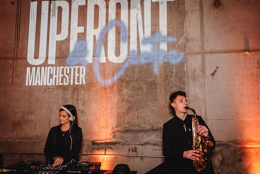 Jamie Anne is positioned to the left, wearing headphones and working the decks. To her right is a saxophonist. Behind them is an industrial concrete wall with signage reading UPFRONT & Centre MANCHESTER